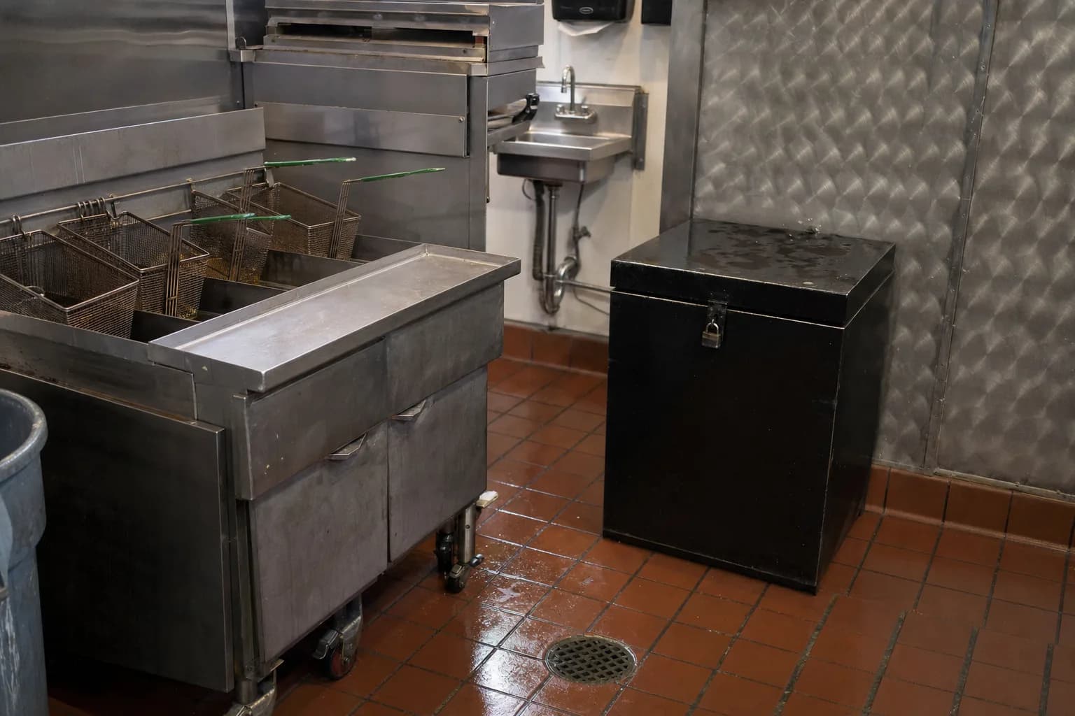 A busy Bay Area commercial kitchen back-of-house with stainless steel fryers, a floor drain and mop sink, and a black locked used cooking oil collection bin against the wall
