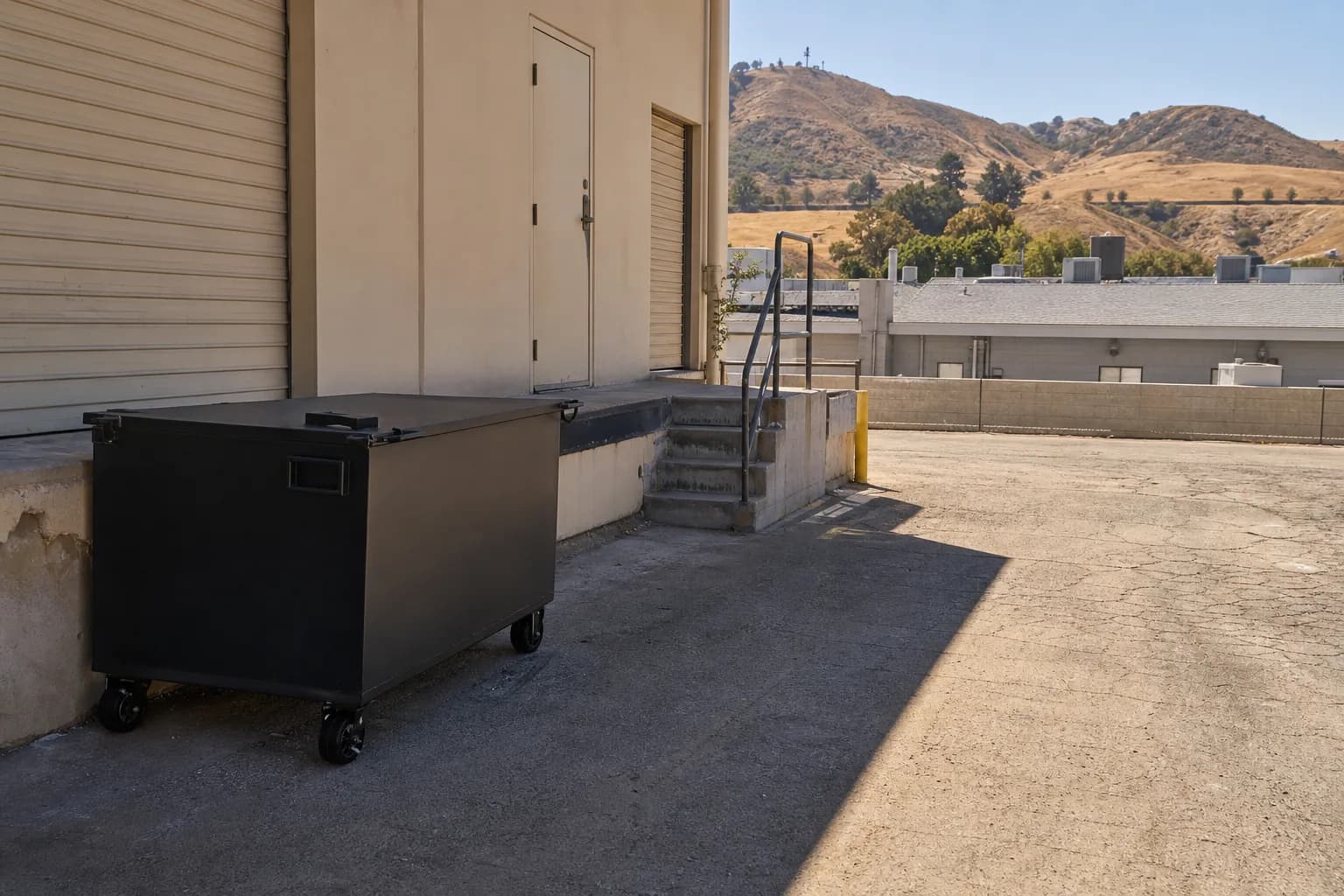 Black locked used cooking oil bin on a shaded concrete pad behind an Inland Empire commercial kitchen, with warehouse rooftops and dry foothills in the background