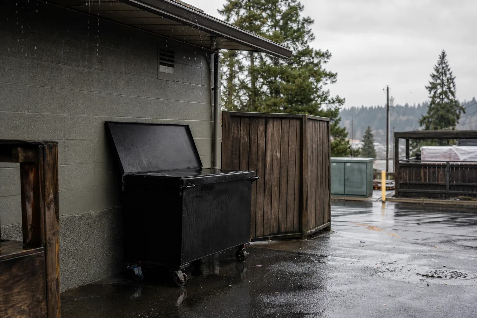 Locked black used cooking oil bin on the rain-damp back dock of a Tacoma restaurant under an overcast South Sound sky, kitchen door propped open behind it