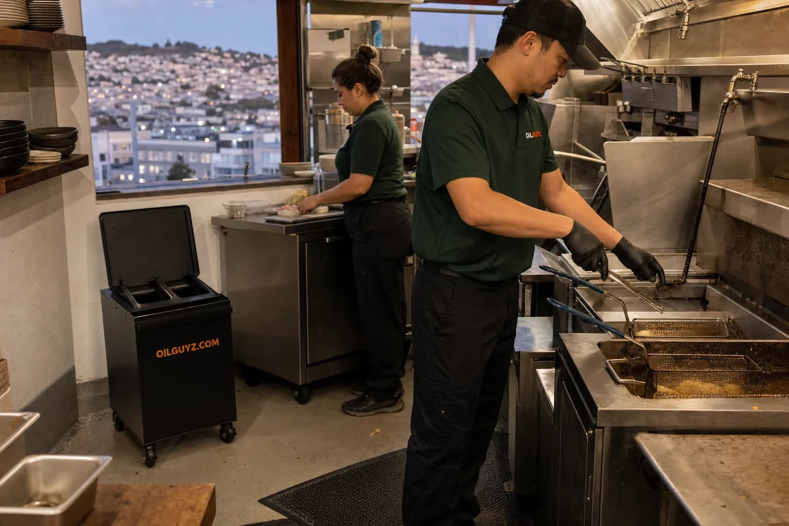 A San Francisco restaurant kitchen line with stainless steel fryers and a grease collection bin, city skyline visible through a window